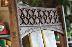 Close-up of the top rail of a wooden chair carved with a repeating scrollwork pattern, with a bookshelf and plant visible blurred in the background