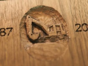 Close-up of a small oval relief carving on wood depicting an excavator, flanked by carved numerals reading '87' on the left and '20' on the right