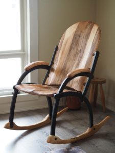 A rocking chair with a live-edge wood seat, backrest, and armrests in light striped tones, joined by black curved metal supports, sitting in a room near a window and small wooden stool.
