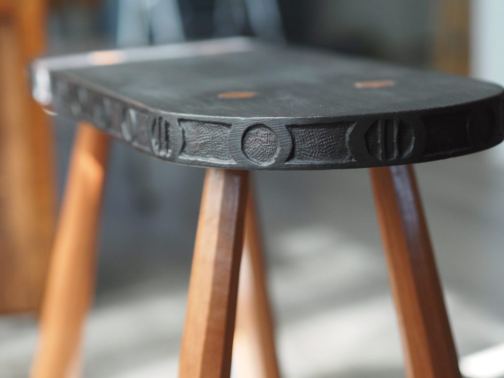 Close-up of a dark carved stool seat edge with alternating ridged and circular relief patterns, supported by light wood tapered legs
